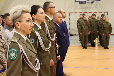 na zdjęciu widoczni zaproszeni goście . na pierwszym planie funkcjonariuszka straży granicznej ubrana w mundur wyjściowy. obok zaproszeni goście z innych formacji mundurowych.  w tle stoją uczniowie klas pierwszych którzy składali ślubowanie.