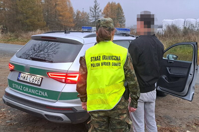 funkcjonariuszka Straży Granicznej ubrana w mundur polo z założoną na siebie kamizelką koloru żółtego z napisem straż graniczna polish border guard. obok niej stoi zatrzymany obywatel Mołdawii. przed nimi znajduje się pojazd służbowy straży granicznej w kolorze srebrnym z sygnałami świetlno dźwiękowymi umiejscowionymi na dachu pojazdu. samochód ma zielone wstawki na karoserii z napisem straż graniczna.