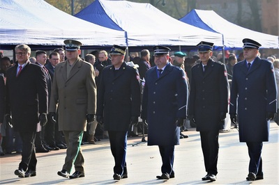 przedstawiciele służb mundurowych idą w jednym szeregu podczas uroczystości. Ida ulicą. Na zdjęciu od strony prawej znajduje się przedstawiciel policji, następnie przedstawiciel straży pożarnej, obok niego idzie również przedstawiciel policji, koło niego również przedstawiciel straży pożarnej, następnie komendant karpackiego oddziału straży granicznej oraz przedstawiciel instytucji państwowej. W tle widoczni są inni uczestniczy uroczystości.