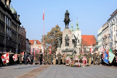 na zdjęciu widoczny grób nieznanego żołnierza. Pod pomnikiem stoją przedstawiciele służb mundurowych którzy tworzą warty honorowe. Przed pomnikiem są złożone kwiaty oraz wiązanki w kolorach białoczerwonych. Z prawej i lewej strony widoczne są sztandary poszczególnych uczestników uroczystości. Obok pomnika, po jego prawej stronie stoi maszt w kolorze białoczerwonym z wciągniętą flagą Polski.