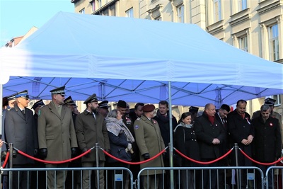 na zdjęciu widoczni uczestnicy uroczystości. Głównie przedstawiciele służb mundurowych. Drugi od lewej stoi komendanta karpackiego oddziału straży granicznej. Osoby znajdują się pod zadaszeniem koloru białego. Przed nimi znajduje się czerwony sznur oddzielający ich od placu uroczystości oraz metalowa barierka.