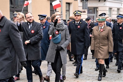 na zdjęciu widoczni uczestnicy przemarszu podczas uroczystości Święta Niepodległości. Na pierwszym planie osoby cywilne trzymające w ręku flagi białoczerwone i machające nimi, natomiast tuż za nimi przedstawiciele służb mundurowych między innymi Zastępca Komendanta Karpackiego Oddziału Straży granicznej – Pani Komendant. Obok niej idzie przedstawiciel straży pożarnej.