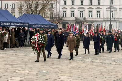 przedstawiciele straży granicznej, straży pożarnej oraz policji idą w kierunku pomnika złożyć wieniec z białoczerwonych kwiatów podczas uroczystości święta niepodległości. Za nimi idą inni uczestnicy uroczystości. Przed nimi idzie funkcjonariusz straży granicznej niosący duży wieniec z kwiatami w kolorze białoczerwonym. W tle widoczne sztandary. Z lewej strony znajdują się zadaszenia w kolorze niebieskim.