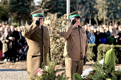 zastępca komendanta karpackiego Oddziału straży granicznej wraz z funkcjonariuszem straży granicznej oddają honor przed pomnikiem podczas uroczystości z okazji święta niepodległości. W tle widoczni inni uczestnicy uroczystości.