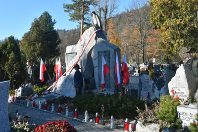 na zdjęciu widoczny pomnik znajdujący się na cmentarzu. pomnik wyryty jest ze skały. znajduje się na nim krzyż a na samej górze wyryty orzeł. obok pomnika umiejscowione są flagi w kolorze białoczerwonym. przed pomnikiem widać zapalone znicze.