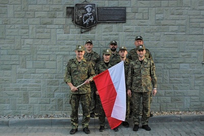 funkcjonariusze straży granicznej stojący pod  ścianą budynku na której zawieszony jest szyld z żołnierzem. jeden z funkcjonariuszy trzyma flagę w kolorze białoczerwonym.