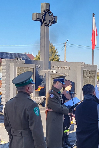 funkcjonariusz straży granicznej odczytujący apel pamięci podczas uroczystości z okazji święta niepodległości. W tle widoczny pomnik. Jest duży krzyż a po jego lewej stronie widać flagę Polski w kolorze biało czerwonym. Z lewej strony zdjęcia stoi również funkcjonariusz straży granicznej.