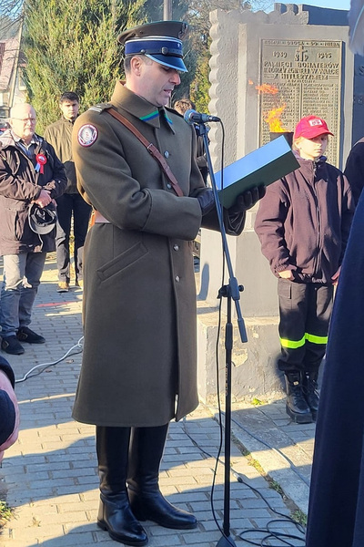 funkcjonariusz straży granicznej odczytujący apel pamięci podczas uroczystości z okazji święta niepodległości. W tle widoczny pomnik oraz inni uczestnicy uroczystości.