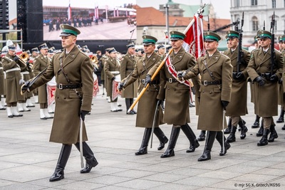 na zdjęciu widoczni funkcjonariusze straży granicznej defilujący podczas uroczystości z okazji święta niepodległości. na czele maszeruje dowódca kompani następnie poczet sztandarowy i reszta funkcjonariuszy idących w szyku i trzymających karabiny.