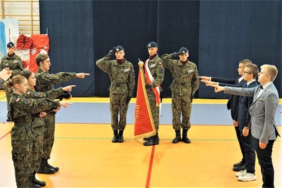 hala widowiskowo- sportowa umundurowani uczniowie stoją po lewej stronie sztandaru a w cywilnych ubraniach po prawej stronie sztandaru. wszyscy trzymają wyciągnięte nad sztandar dłonie i ślubują na sztandar trzymany przez poczet sztandarowy