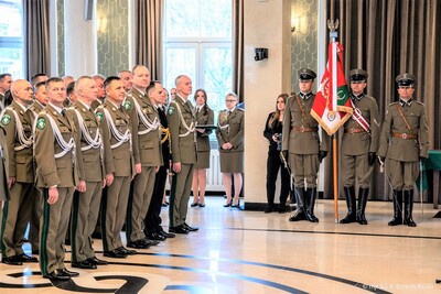 na pierwszym planie widoczni komendanci wszystkich oddziałów straży granicznej. po prawej ich stronie stoi poczet sztandarowy wraz ze sztandarem straży Granicznej. uroczystość odbywa się w pomieszczeniu komendy głównej straży granicznej.