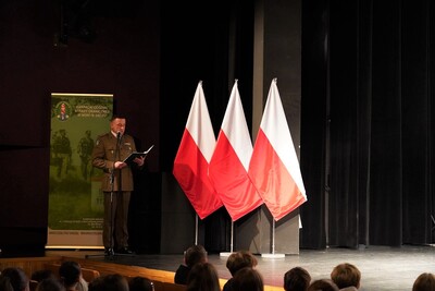 na zdjęciu widoczny na scenie kina funkcjonariusz straży granicznej, który prowadzi całe wydarzenie. obok niego położone są trzy białoczerwone flagi Polski.