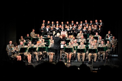 na zdjęciu widoczna orkiestra reprezentacyjna straży granicznej oraz dyrygujący nią jubilat Bolesław Szulia ubrany w czarny garnitur. na dalszym planie widoczny chór. koncert odbywa się na scenie w pomieszczeniu.