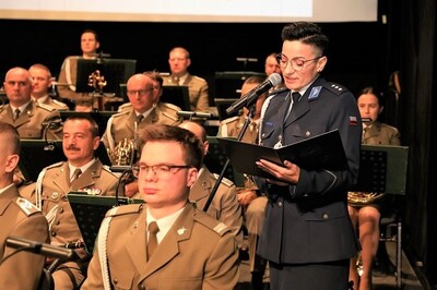 Na tle siedzącej na scenie orkiestry policjantka wyczytująca wyróżnionych i odznaczanych policjantów.