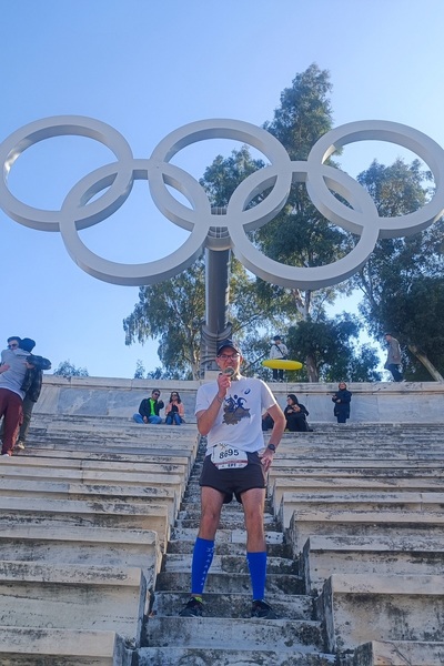 bohater artykułu stoi na schodach stadionu. w rękach trzyma medal. w tle pięć kół olimpijskich w kolorze jasnoszarym na postumencie