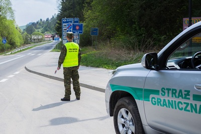 funkcjonariusz straży granicznej patrolujący na ulicy. za nim postawiony jest samochód służbowy straży granicznej z napisami na drzwiach straż graniczna.