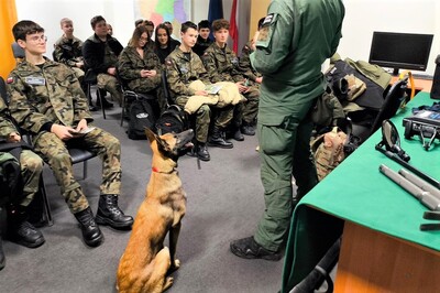 uczniowie siedzą na krzesłach, przed nimi pies służbowy siedzi wpatrzony w przewodnika, który opowiada uczniom o swojej służbie,  na stole leży sprzęt służbowy wykorzystywany na co dzień w służbie