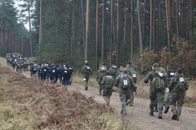 grupa kilkudziesięciu osób w mundurach zielonych i granatowych biegnie przez teren leśny . jest jesień, na podłożu dużo liści, teren nierówny