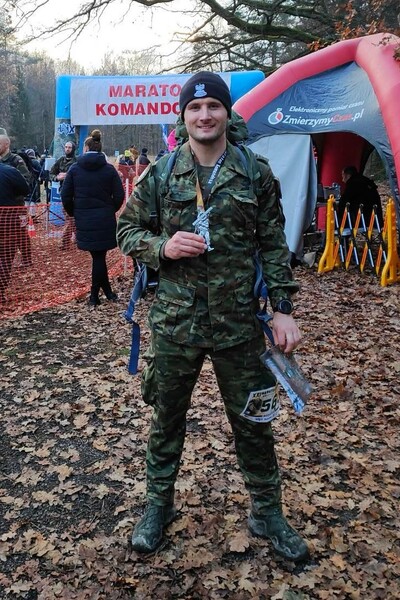 funkcjonariusz kaosg stoi z medalem ukończenia maratonu komandosa w ręku. jest trochę wybrudzony błotem, ubrany w mundur i wojskowe buty oraz czapkę czarna bawełniana z orzełkiem, w tle meta biegu i inni uczestnicy