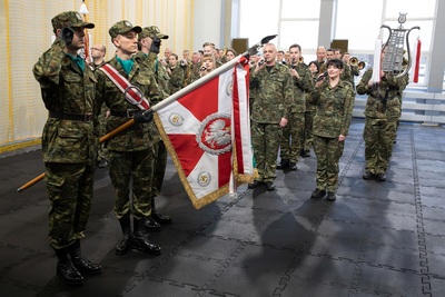 poczet sztandarowy wraz ze sztandarem karpackiego oddziału straży granicznej prezentuje sztandar. za pocztem stoją muzycy orkiestry reprezentacyjnej straży granicznej wykonujący hymn Polski. na sobie mają umundurowanie polo. uroczystość odbywa się w hali sportowej.