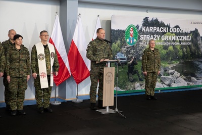 komendant karpackiego oddziału straży granicznej przemawia do wszystkich zebranych podczas uroczystości. przed sobą ma mikrofon oraz mównice. za nim widoczne są flagi białoczerwone oraz baner promocyjny oddziału z napisem karpacki oddział straży granicznej. tuz za komendantem po jego lewej stroni stoi jego zastępca, pani komendant a po jego prawej stronie stoi kapelan oddziału oraz widoczna jest część kadry kierowniczej oddziału. wszyscy maja na sobie mundur polo. uroczystość odbywa się w hali sportowej.