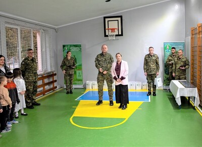 sala gimnastyczna zespołu szkolno-przedszkolnego nr 7 w nowym sączu. pani dyrektor mówi do zgromadzonych dzieci, które brały udział w konkursie. Obok niej stoi generał Jopek a trochę z tyłu jedna funkcjonariuszka i czterech funkcjonariuszy. Jeden stoi pod oknem, funkcjonariuszka trzyma teczkę otwartą z czytanym tekstem, jeden trzyma torebki z nagrodami.