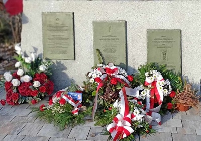 na zdjęciu widoczny jest obelisk upamiętniający wydarzenia z 1944 roku . pod obeliskiem złożone są wiązanki kwiatów przyozdobione bało czerwonymi wstęgami.