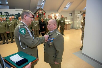 kapelan karpackiego oddziału straży granicznej przypina medal od biskupa polowego wojska polowego funkcjonariuszowi straży granicznej, który odchodzi na emeryturę. Na dalszym planie widoczni są funkcjonariusze oraz pracownik Karpackiego Oddziału straży granicznej. Uroczystość odbywa się w Sali tradycji Karpackiego Oddziału Straży Granicznej.