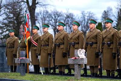 Na zdjęciu widać mężczyzn ubranych w mundury, którzy stoją w szeregu, jeden trzyma sztandar. Z przodu widać dwa kamienne krzyże wbite w zieloną trawę, na jednym położony jest biały aniołek z gipsu. Krzyże przewiązane są biało-czerwonymi wstążkami, po lewej stronie zdjęcia widać dużą czarną tablicę z napisami w kolorze białym.