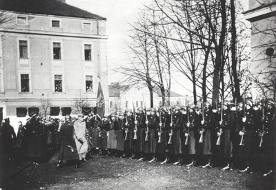 zdjęcie ukazuje fotografie z czasów między wojennych na której widoczny jest marszałek Piłsudski który wizytował koszary w nowym Sączu. w tle widoczne są budynki koszar. przed marszałkiem stoi kompania honorowa złożona z żołnierzy.