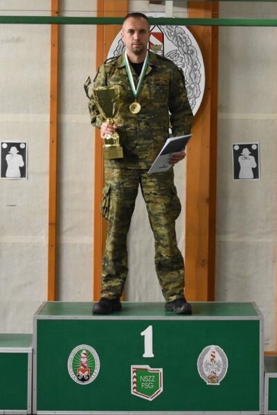 Funkcjonariusz sg stoi na 1 stopniu podium. w rękach trzyma puchar i dyplom, a na szyi ma zawieszony medal.