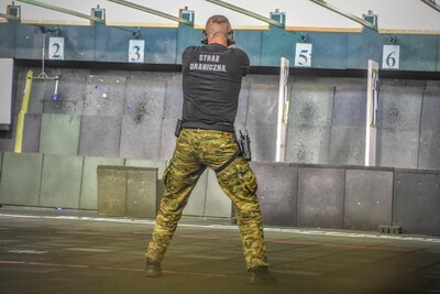 funkcjonariusz w słuchawkach zagłuszających na uszach strzela z broni krótkiej do obrotowych tarcz. Ujęcie z tyłu.