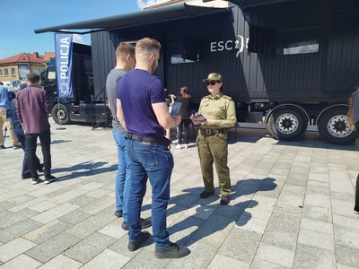 Na zdjęciu widać grupę ludzi, w tle stoi dużą ciężarówkę na której stoi kontener w kolorze czarnym. Z boku widać niebieski napis policja.