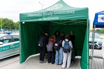 Namiot w kolorze zielonym z napisem Placówka Straży Granicznej w Tarnowie. Grupa młodzieży stoi tyłem i słucha dwóch kobiet w mundurze. Z prawej strony widać samochód służbowy srebrny z zielonym napisem Straż Graniczna.