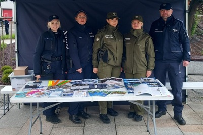 Na zdjęciu stoi grupa pięciu osób ubrana w mundury Policji i Straży Granicznej na tle granatowego namiotu. Przed nimi stoi stół z wieloma ulotkami promocyjnymi. Stół jest w kolorze białym na metalowych nogach w kolorze popielatym.