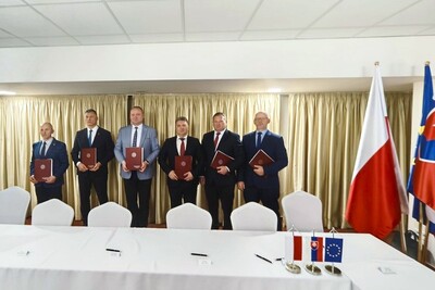 Na zdjęciu widać sześciu mężczyzn stojących pod ścianą. W rękach trzymają bordową teczkę na dokumenty na której jest logo. Z prawej strony widać flagi : Polski jest biało czerwona, a druga Słowacji niebiesko czerwona z logiem na środku. Z przodu zdjęcia widać stół na którym są proporczyki z flagami Polski i Słowacji oraz Unii Europejskiej. Na stole leżą czarne długopisy.