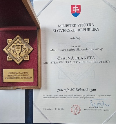Po lewej stronie zdjęcia widnieje złote odznaczenie w bordowej szkatułce z napisami w języku słowackim po prawej stronie jest zaświadczenie w kolorze białym o odznaczeniu napisane w języku słowackim z herbem Słowacji na górze zaświadczenia cale zaświadczenie jest  nadrukowane na białym papierze ze znakiem wodnym przypominającym odznaczenie