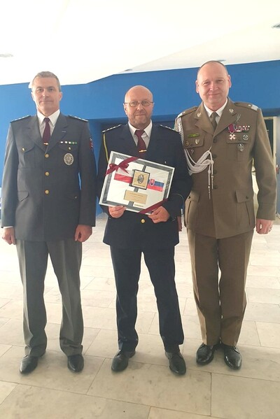 Na zdjęciu 3 osoby w mundurach jeden funkcjonariusz Straży Granicznej 2 pozostałych zagraniczni mundurowi, ubrani w mundury wyjściowe mężczyzna na środku trzyma w rękach odznaczenie z flagami Polski i Słowacji
