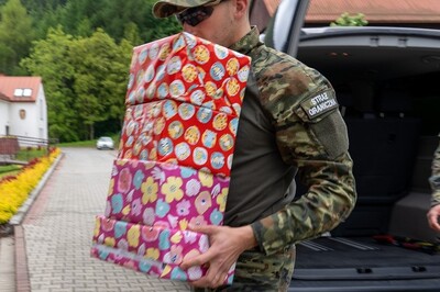 Funkcjonariusz ubrany w mundur polowy, niesie prezenty zapakowane w kolorowe pudełka na ramieniu ma naszywkę z napisem straż graniczna