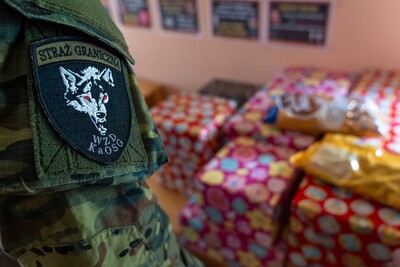 Na pierwszym planie widać ramie funkcjonariusza straży granicznej  z  naszywką straży granicznej wydziały zabezpieczenia działań z charakterystycznym wilkiem, w tle widać prezenty ułożone na stole