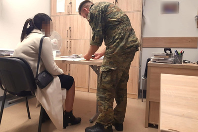 Na zdjęciu widać kobietę i mężczyznę w pokoju. Kobieta siedzi na krześle, ubrana w jasny płaszcz, czarną spódnicę i czarne buty. Na ramieniu ma czarną małą torebkę. Mężczyzna ubrany jest w zielony mundur oraz czarne buty. Na zdjęciu widać meble w jasnym kolorze. Mężczyzna przedstawia kobiecie dokumenty do podpisu.