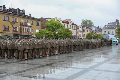 Grupa ludzi ubrana w mundury stoi na płycie rynku. Płyty są mokre od deszczu. W tle stoi rząd budynków. Widoczne są zielone drzewa. Z prawej strony widać duży samochód w kolorze popielatym.