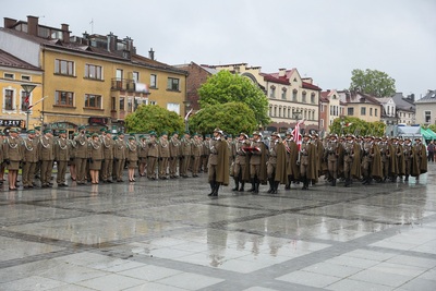Grupa ludzi ubrana w mundury stoi na płycie rynku. Kolejna grupa maszeruje środkiem. Płyty są mokre od deszczu. W tle stoi rząd budynków. Widoczne są zielone drzewa. Z prawej strony widać duży samochód w kolorze popielatym.