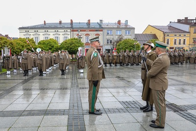 Trzech funkcjonariuszy stoi na froncie zdjęcia, dwóch składa meldunek. W tle widać grupę osób ubraną w mundury, stoją w rzędzie. W tle stoi rząd budynków. Widoczne są zielone drzewa. Z prawej strony widać duży samochód w kolorze popielatym.
