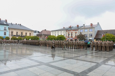 Grupa ludzi ubrana w mundury stoi na płycie rynku. Dwie osoby maszerują środkiem. Płyty są mokre od deszczu. W tle stoi rząd budynków, między drzewami powiewają flagi biało-czerwone. Widoczne są zielone drzewa.