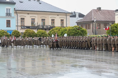 Grupa ludzi ubrana w mundury stoi na płycie rynku. Trzy osoby maszerują środkiem, jedna trzyma w rękach złożoną flagę biało-czerwoną. Płyty są mokre od deszczu. W tle stoi rząd budynków, między drzewami powiewają flagi biało-czerwone. Widoczne są zielone drzewa.