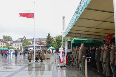 Na płycie rynku stoją osoby. Trzy osoby stoją przy maszcie koloru popielatego na którym powiewa biało-czerwona flaga. Z prawej strony widać namiot w kolorze zielonym z sufitem w kolorze beżowym. Pod namiotem stoją osoby, które patrzą na flagę.