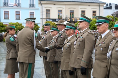 Na zdjęciu widać sześć osób, które ubrane są w mundury. Jedna osoba gratuluje podając rękę osobie z kapeluszem z piórkiem na głowie. W tle stoi rząd budynków. Widoczne są zielone drzewa.