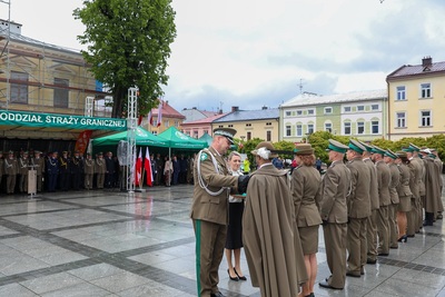 Na zdjęciu widać piętnaście osób, które ubrane są w mundury, jedna ubrana jest w czarną spódnicę i biały żakiet. Mężczyzna przypina drugiemu mężczyźnie w mundurze z peleryną i kapeluszem na głowie medal. Z lewej strony zdjęcia widać cztery zielone namioty pod którymi stoi wiele osób. Na środku widać flagi biało-czerwone. W tle stoi rząd budynków. Widoczne są zielone drzewa. Płyty są mokre od deszczu.