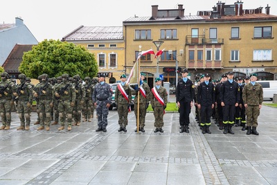 Grupa ludzi ubrana w mundury stoi na płycie rynku. Płyty są mokre od deszczu. W tle stoi rząd budynków. Widoczne są zielone drzewa. Na środku stoją trzy osoby , które mają przepasane biało-czerwone szarfy na piersi. Jedna osoba trzyma w rękach sztandar. W tle powiewają dwie flagi biało czerwone. W tle stoi rząd budynków. Widoczne są zielone drzewa.